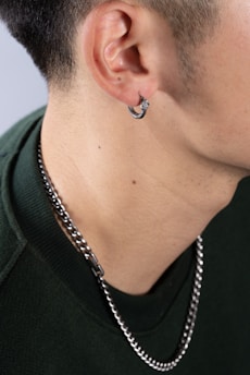 A close-up of a person's neck and ear, showing a silver hoop earring in the ear and a silver chain necklace around the neck. The person is wearing a green garment.