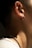 A close-up of a person's ear and neck is visible, featuring an earring on the ear and a chain necklace around the neck. The lighting creates soft shadows, highlighting the texture of the skin and jewelry.