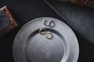 A matte metal plate is placed on a dark surface, with pairs of textured metal hoop earrings resting on it. The earrings are in different colors, including gold and silver. A wooden box with metal details is partially visible in the upper left corner.