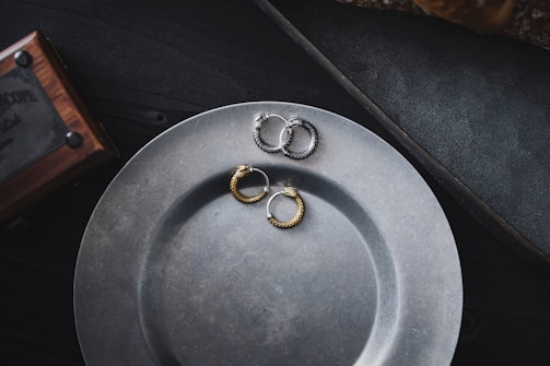 A matte metal plate is placed on a dark surface, with pairs of textured metal hoop earrings resting on it. The earrings are in different colors, including gold and silver. A wooden box with metal details is partially visible in the upper left corner.