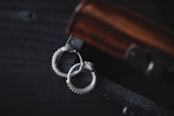 A pair of intricately designed silver earrings with a circular, snake-like motif lies gracefully on a dark wooden surface. The earrings exhibit detailed craftsmanship with a textured, scaly pattern, adding to their elegant appearance. The wooden surface has a rich, warm tone with visible grain patterns, complementing the metallic sheen of the earrings.
