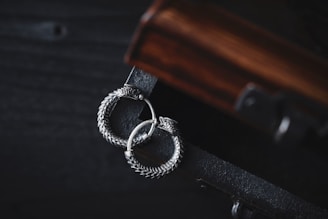A pair of intricately designed silver earrings with a circular, snake-like motif lies gracefully on a dark wooden surface. The earrings exhibit detailed craftsmanship with a textured, scaly pattern, adding to their elegant appearance. The wooden surface has a rich, warm tone with visible grain patterns, complementing the metallic sheen of the earrings.