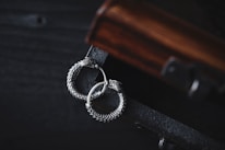 A pair of intricately designed silver earrings with a circular, snake-like motif lies gracefully on a dark wooden surface. The earrings exhibit detailed craftsmanship with a textured, scaly pattern, adding to their elegant appearance. The wooden surface has a rich, warm tone with visible grain patterns, complementing the metallic sheen of the earrings.