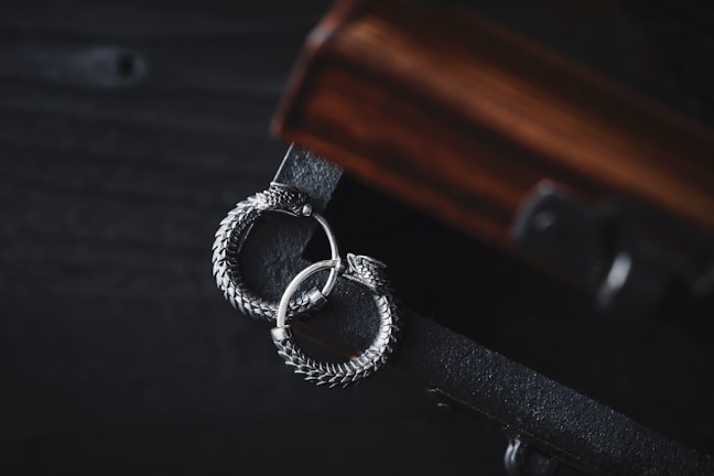 A pair of intricately designed silver earrings with a circular, snake-like motif lies gracefully on a dark wooden surface. The earrings exhibit detailed craftsmanship with a textured, scaly pattern, adding to their elegant appearance. The wooden surface has a rich, warm tone with visible grain patterns, complementing the metallic sheen of the earrings.