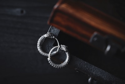 A pair of intricately designed silver earrings with a circular, snake-like motif lies gracefully on a dark wooden surface. The earrings exhibit detailed craftsmanship with a textured, scaly pattern, adding to their elegant appearance. The wooden surface has a rich, warm tone with visible grain patterns, complementing the metallic sheen of the earrings.
