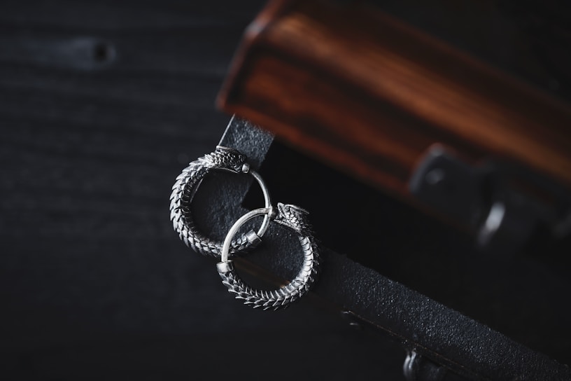 A pair of intricately designed silver earrings with a circular, snake-like motif lies gracefully on a dark wooden surface. The earrings exhibit detailed craftsmanship with a textured, scaly pattern, adding to their elegant appearance. The wooden surface has a rich, warm tone with visible grain patterns, complementing the metallic sheen of the earrings.