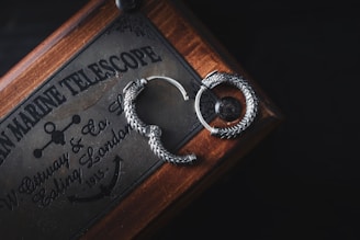 A pair of intricately designed silver earrings shaped like serpents lie on a wooden box. The box has vintage inscriptions and markings, indicating a historical or nautical theme, with references to a marine telescope and a company in London. The scene is lit softly, highlighting the metallic details and craftsmanship of the earrings against the dark background.