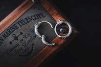A pair of intricately designed silver earrings shaped like serpents lie on a wooden box. The box has vintage inscriptions and markings, indicating a historical or nautical theme, with references to a marine telescope and a company in London. The scene is lit softly, highlighting the metallic details and craftsmanship of the earrings against the dark background.