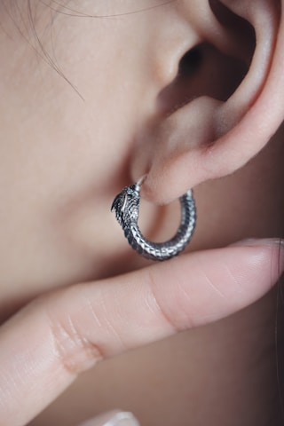 A close-up of an ear adorned with a silver earring shaped like a coiled serpent. The intricate design of the earring suggests it is crafted with attention to detail. A finger is gently touching the lower part of the earlobe, drawing attention to the earring.