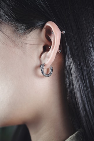 A close-up of a person's ear adorned with multiple piercings. The ear is decorated with a silver hoop earring in the earlobe, and additional studs in the cartilage. The person's hair is dark, straight, and tucked behind the ear, and the skin tone is light.
