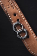 Two intricately designed silver earrings with a serpentine motif rest on a textured brown leather strap. The strap features detailed stitching and embossed text, partially displaying the words 'DREAM STAR DESIGN 24'. The earrings appear to be circular with detailed scales, resembling intertwined serpents.