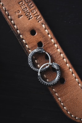 Two intricately designed silver earrings with a serpentine motif rest on a textured brown leather strap. The strap features detailed stitching and embossed text, partially displaying the words 'DREAM STAR DESIGN 24'. The earrings appear to be circular with detailed scales, resembling intertwined serpents.