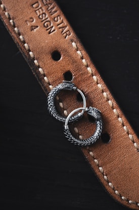 Two intricately designed silver earrings with a serpentine motif rest on a textured brown leather strap. The strap features detailed stitching and embossed text, partially displaying the words 'DREAM STAR DESIGN 24'. The earrings appear to be circular with detailed scales, resembling intertwined serpents.