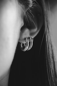 A close-up of a person's ear adorned with a textured, metallic hoop earring. The photograph is captured in black and white, emphasizing contrasts and details. The person's hair flows down, partially framing the ear.