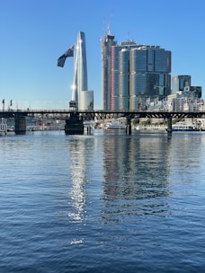 A modern cityscape featuring a prominent glass skyscraper and a row of other tall buildings under a clear blue sky. The foreground shows a calm body of water reflecting the buildings and bridge above it. A large flag is visible near the center, and cranes can be seen atop the buildings, suggesting construction activity.