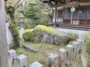 A tranquil garden scene featuring a large moss-covered rock surrounded by a low stone fence. The garden includes well-trimmed bushes and a tree with gnarled branches. In the background, there is a traditional wooden building with sliding doors and a hanging lantern.