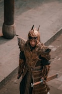 A person is wearing ornate, bronze-colored armor with intricate designs, standing on a stone pavement. The armor appears to be of a historical or fantasy design, including a helmet with pointed spikes and a long sword held in one hand. The setting suggests an ancient or medieval theme.