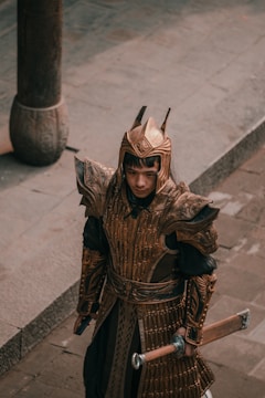 A person is wearing ornate, bronze-colored armor with intricate designs, standing on a stone pavement. The armor appears to be of a historical or fantasy design, including a helmet with pointed spikes and a long sword held in one hand. The setting suggests an ancient or medieval theme.