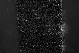 A close-up view of an array of small, circular, hollow tubes densely packed together. The tubes are various sizes and appear to be made of a dark, possibly metallic material. Some areas have a web-like substance partially covering the tubes, giving the image an organic, almost eerie feel.