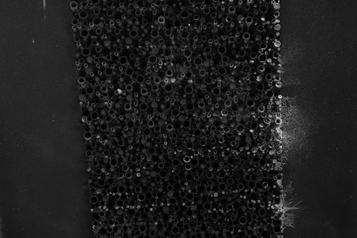 A close-up view of an array of small, circular, hollow tubes densely packed together. The tubes are various sizes and appear to be made of a dark, possibly metallic material. Some areas have a web-like substance partially covering the tubes, giving the image an organic, almost eerie feel.
