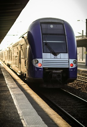 A blue and gray passenger train is approaching a platform, bathed in warm sunlight. The train is on a railway track at a train station, with the station's platform visible on the left side of the image. The early morning or late afternoon light casts long shadows, and there are overhead power lines in the background.