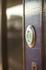 A close-up view of a modern electronic door lock with a circular design, featuring a green illuminated button. The lock is installed on a wooden surface, and the background is softly blurred.