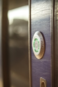 A close-up view of a modern electronic door lock with a circular design, featuring a green illuminated button. The lock is installed on a wooden surface, and the background is softly blurred.