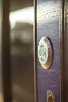 A close-up view of a modern electronic door lock with a circular design, featuring a green illuminated button. The lock is installed on a wooden surface, and the background is softly blurred.