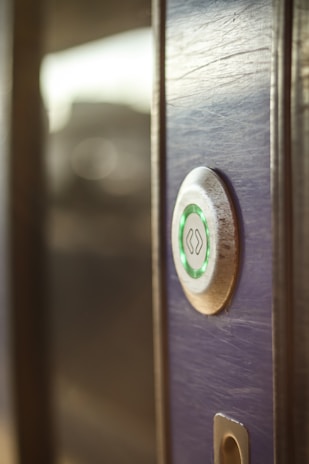 A close-up view of a modern electronic door lock with a circular design, featuring a green illuminated button. The lock is installed on a wooden surface, and the background is softly blurred.