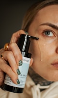 A person is applying a liquid cleanser to their face using a bottle with a pump. The focus is on the person's eye and hand, which has painted nails and a ring.