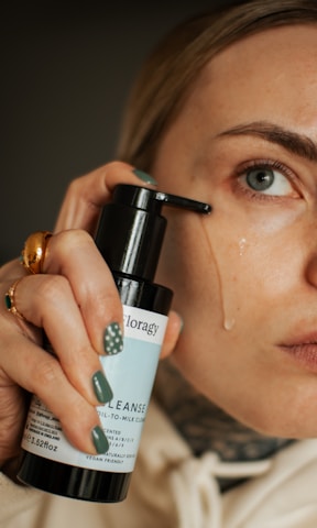 A person is applying a liquid cleanser to their face using a bottle with a pump. The focus is on the person's eye and hand, which has painted nails and a ring.