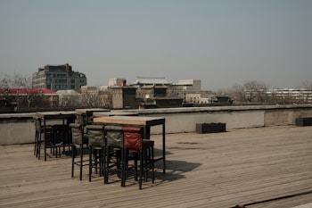 A rooftop area with wooden flooring featuring a set of high tables and chairs. The background showcases an urban landscape with a mix of modern and traditional architecture, with buildings and leafless trees visible under a clear sky.
