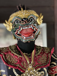 An intricately designed traditional mask featuring a fierce expression with wide eyes, an open mouth, and detailed carvings. The mask is adorned with golden decorations, and the garment has ornate patterns in red and gold.