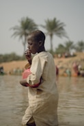 A person in a light-colored garment stands in shallow water, holding a red plastic container. Palm trees can be seen in the blurred background, along with other people in the distance.