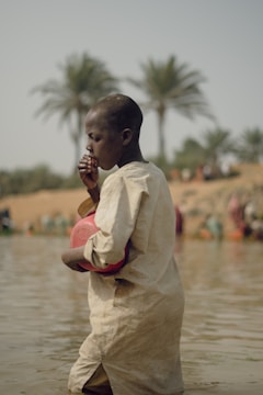 A person in a light-colored garment stands in shallow water, holding a red plastic container. Palm trees can be seen in the blurred background, along with other people in the distance.