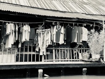 A collection of clothes hangs on a line outside a rustic wooden house with a corrugated metal roof. A clothes drying rack is also visible, along with potted plants placed on the porch. A bicycle can be seen behind the clothes, partially obscured. The scene appears calm and everyday, suggesting domestic life by the waterfront.
