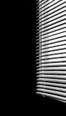Vertical blinds partially covering a window, creating striped patterns of light and shadow across a dimly lit space. The contrast emphasizes the division between the illuminated and dark areas.