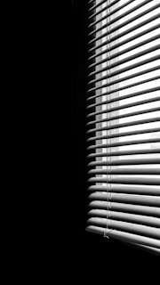 Vertical blinds partially covering a window, creating striped patterns of light and shadow across a dimly lit space. The contrast emphasizes the division between the illuminated and dark areas.