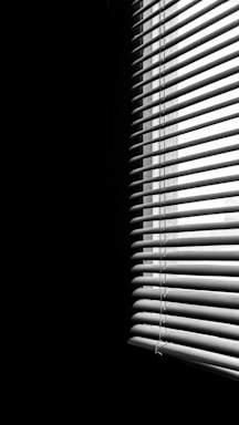 Vertical blinds partially covering a window, creating striped patterns of light and shadow across a dimly lit space. The contrast emphasizes the division between the illuminated and dark areas.