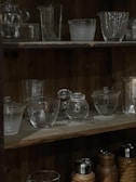 A wooden shelf displaying various clear glassware and bottles with a dimly lit ambiance. The top shelf holds a variety of glass containers including cups and jugs, showcasing different textures and shapes. The bottom shelf has both glass and metal containers, with some covered in a woven material.