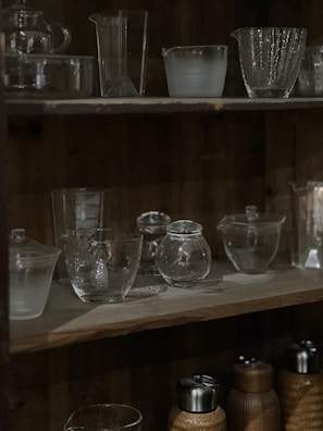 A wooden shelf displaying various clear glassware and bottles with a dimly lit ambiance. The top shelf holds a variety of glass containers including cups and jugs, showcasing different textures and shapes. The bottom shelf has both glass and metal containers, with some covered in a woven material.