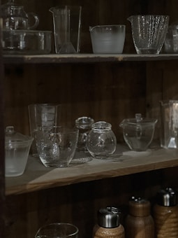 A wooden shelf displaying various clear glassware and bottles with a dimly lit ambiance. The top shelf holds a variety of glass containers including cups and jugs, showcasing different textures and shapes. The bottom shelf has both glass and metal containers, with some covered in a woven material.