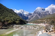 A scenic landscape featuring a tiered water terrace with clear, turquoise pools set against a backdrop of majestic snow-capped mountains and lush, green wooded slopes. People are visible walking along a pathway and gathering near the water's edge under a clear blue sky.