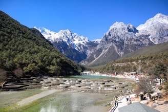 A scenic landscape featuring a tiered water terrace with clear, turquoise pools set against a backdrop of majestic snow-capped mountains and lush, green wooded slopes. People are visible walking along a pathway and gathering near the water's edge under a clear blue sky.