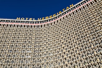A large architectural structure with a detailed facade composed of a repeating pattern of square and rectangular windows. The building has a prominent signage at the top in bold letters spelling 'HOTEL UZBEKISTAN' against a clear blue sky.