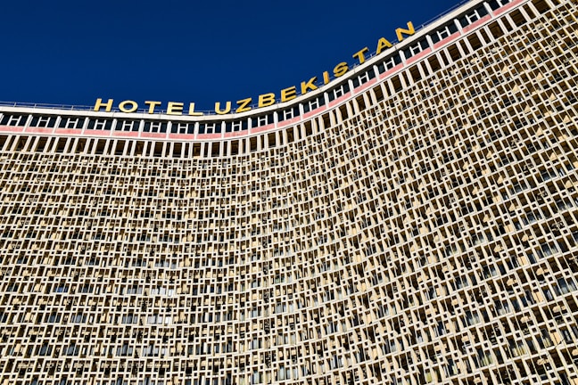 A large architectural structure with a detailed facade composed of a repeating pattern of square and rectangular windows. The building has a prominent signage at the top in bold letters spelling 'HOTEL UZBEKISTAN' against a clear blue sky.