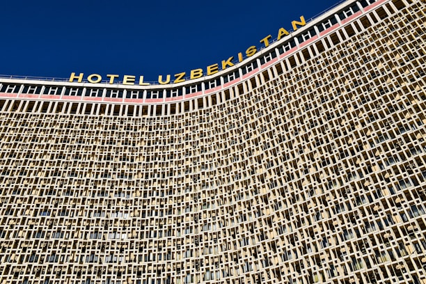 A large architectural structure with a detailed facade composed of a repeating pattern of square and rectangular windows. The building has a prominent signage at the top in bold letters spelling 'HOTEL UZBEKISTAN' against a clear blue sky.