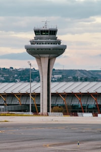 A tall, cylindrical airport control tower stands prominently near a terminal building with a curved roof. In the background, low hills and scattered buildings are visible under a cloudy sky.
