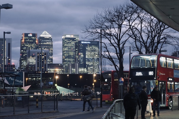 A cityscape featuring modern skyscrapers illuminated at dusk, with prominent signs of financial institutions. In the foreground, a red double-decker bus with lights on is stationary, and several pedestrians are visible. Leafless trees add contrast to the scene, and there are clear signs directing towards a district.