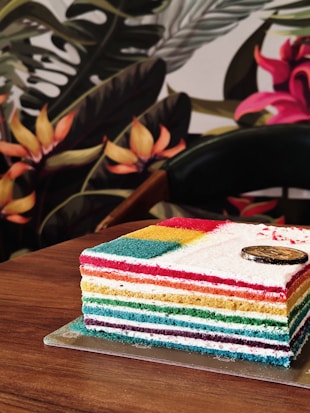 A multi-layer rainbow cake featuring vibrant layers in red, orange, yellow, green, blue, and purple, topped with a small circular decoration. The cake rests on a wooden table, set against a background of colorful and lush tropical foliage, including flowers and large leaves.